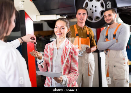 Client at a car dealership giving keys from her car to vehicle repair crew manager. Confident smiling mechanics are standing in a background Stock Photo
