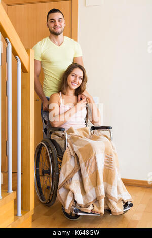 Young positive man and his pretty disabled wife in wheelchair coming home. Focus on woman Stock Photo