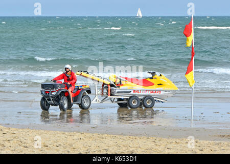 RNLI Lifeguard on the shoreline at Sandbanks beach driving a ATV quad bike and Yamaha jet ski PWC rescue craft with choppy sea & sailing boat distant Stock Photo