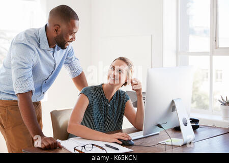 Man stands talking to woman smiling at her desk in an office Stock Photo