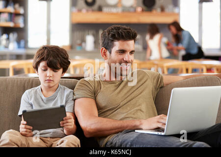 Father And Son Sitting On Lounge Sofa Using Digital Devices Stock Photo