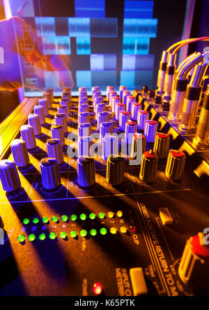 Close-up of an audio mixing desk with background computer screen. Motion blurred hand on distant controls. Selective focus on mixer controls Stock Photo