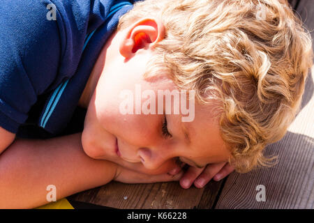 Child, little blonde curly haired boy, close up of face, pretending to be asleep, head resting on hands while laying on wooden deck. Stock Photo