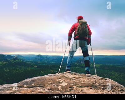 Disabled man with crutches stands on a big rock and looking to mountains at horizon. Hiker silhouette with medicine crutch on mountain peak.