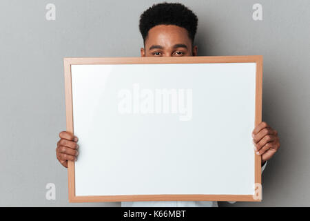 Young afro american man hiding behind blank board isolated over gray background Stock Photo