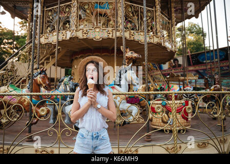 Young beautiful girl eating ice cream while standing in front of the carousel at the amusement park Stock Photo