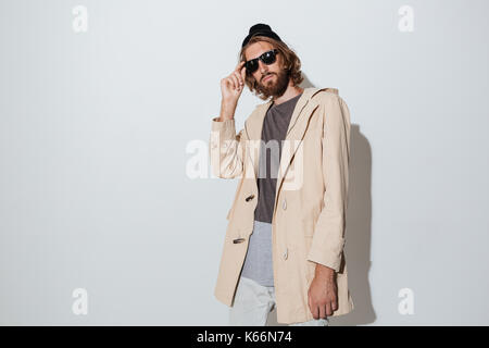 Picture of young bearded hipster man wearing sunglasses standing isolated over grey wall. Looking camera. Stock Photo