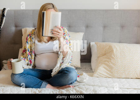 Caucasian expectant mother sitting on bed reading book and drinking coffee Stock Photo