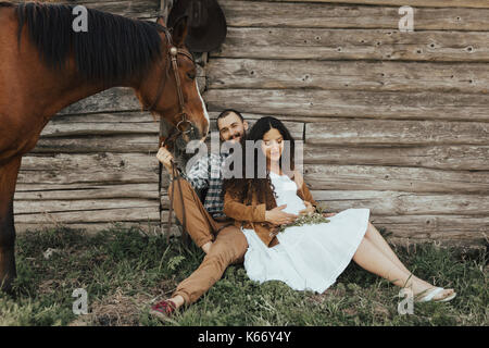 Couple sitting in grass near barn with horse Stock Photo
