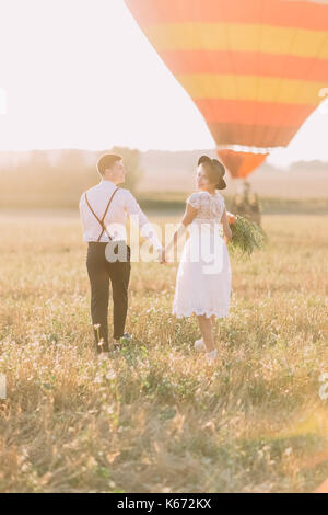 The back view of the vintage dressed newlyweds walking in the field near the airballoon. Stock Photo