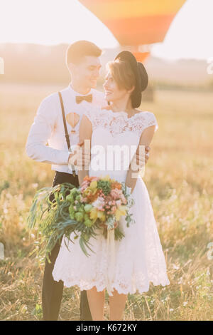 The happy groom is hugging thesmiling bride with the colourful bouquet back in the sunny field during the sunset. Stock Photo