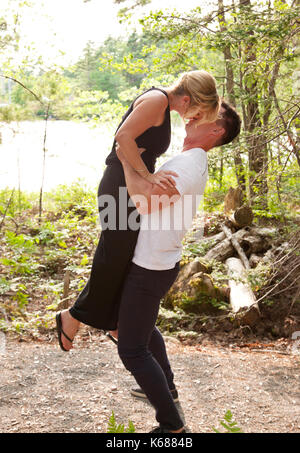 a strong handsome man lifts a beautiful blond woman in the air while they kiss in the forest Stock Photo