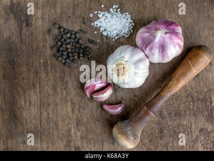 Top view of spices and mortar and pestle, showing whole purple garlic bulbs, garlic cloves, black pepper and white salt, on top of a wooden surface Stock Photo