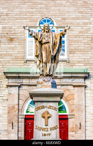 Ile D'Orleans, Canada - June 1, 2017: Saint-Jean red painted church with stone architecture and Jesus Christ statue with outstretched open welcome arm Stock Photo