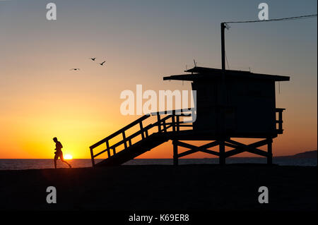 Silhouettes of a sportive runner working out on the sand and the iconic lifeguard tower with flying birds at sunset in Venice Beach, CA. Stock Photo