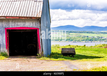 Blue and red painted old vintage shed or barn with wooden wagon overlooking river in summer landscape field in countryside Stock Photo