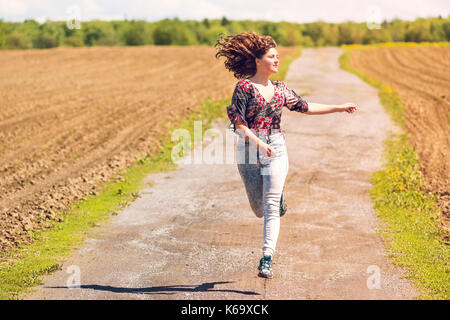 Young woman running, jumping in air and smiling on countryside dirt road by brown plowed fields with furrows in summer in Ile D'Orleans, Quebec, Canad