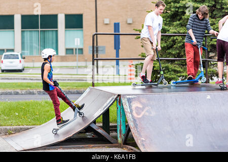 Saguenay, Canada - June 3, 2017: Downtown city summer park in Quebec with young teenagers boys skating, performing tailwhip tricks, stunts on scooter  Stock Photo