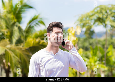 Portrait Of Happy Smiling Man Making Phone Call Over Tropical Forest And Blue Sky Background Stock Photo