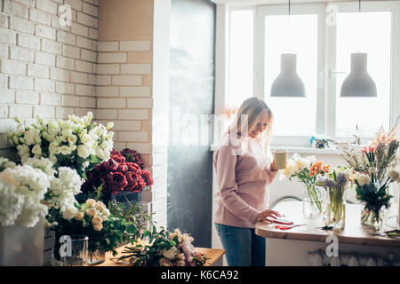 Florist at work. A woman in the process of making a summer bouquet of peonies, holds in her hand a cup of coffee. Tools and accessories florists need  Stock Photo