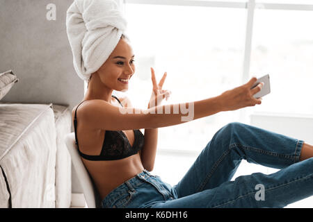 Smiling young asian woman with bath towel wrapped around her head taking a selfie while sitting on a chair at home Stock Photo