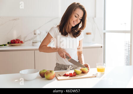 Smiling young woman cutting fruits on a wooden board while making breakfast in a kitchen Stock Photo