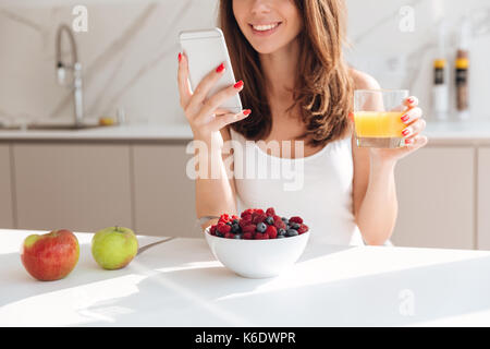 Cropped image of smiling pretty woman looking at mobile phone and holding glass of orange juice while having breakfast in a kitchen Stock Photo
