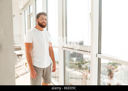 Smiling pensive bearded man standing near the window at home Stock Photo