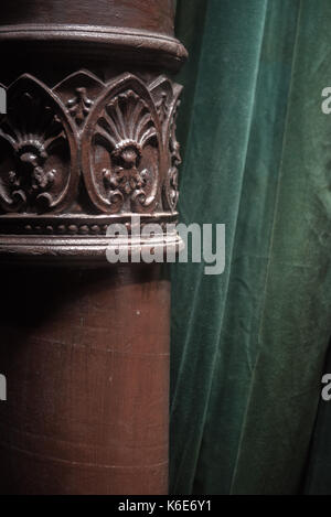 Ornate acanthus decoration on a brown painted cast iron pole in a room interior in front of a green drape or curtain Stock Photo