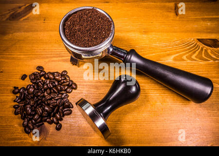 A handful of coffee beans, alongside ground coffee in an espresso machine portafilter basket and a steel tamper, all sitting on a pine worktop. Stock Photo