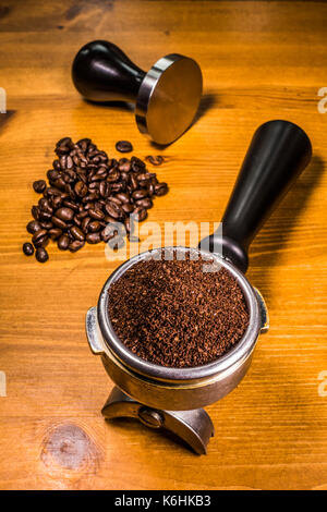 A handful of coffee beans, alongside ground coffee in an espresso machine portafilter basket and a steel tamper, all sitting on a pine worktop. Stock Photo