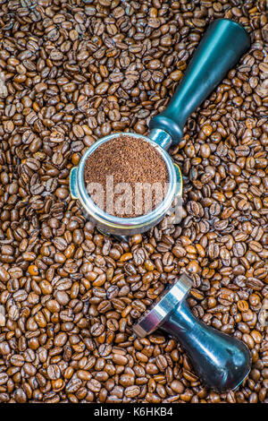 Espresso making concept: Ground coffee in an espresso machine portafilter basket, with steel tamper alongside, on top of lots of coffee beans. Stock Photo