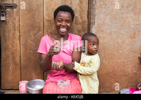 Lugazi, Uganda. 09 June 2017. A laughing African mother with a little child boy. The woman is sitting on door threshold sorting grains. Stock Photo