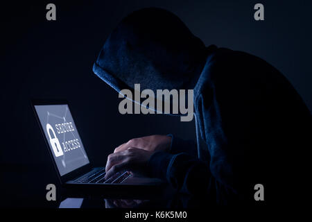 Hooded hacker in dark room performing a cyber attack to a secure access to steal data on internet, laptop computer screen Stock Photo
