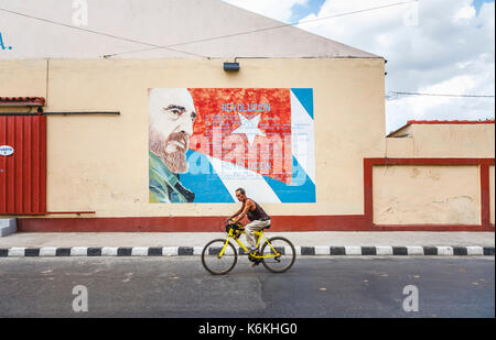 Local man riding a bicycle cycling past a roadside wall painting of Fidel Castro and the Cuban flag, Cienfuegos, a city on the south coast of Cuba Stock Photo