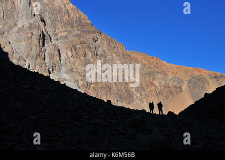 Morocco, North Africa, High Atlas Mountains. Two friends trekking on the rocky mountain path against the rocks and blue sky. Stock Photo