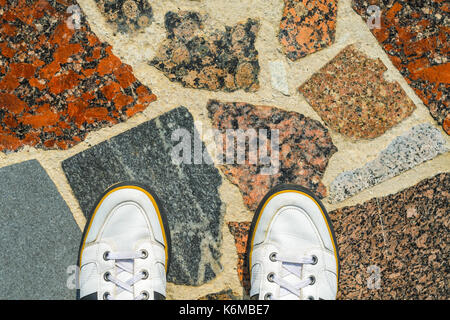 White Sneakers on a Multi-Material Mosaic Paving Stone Made of Granite, Marble, Natural Stone Tiles and Concrete Top View. Travel, Lifestyle Concept.  Stock Photo