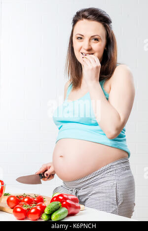 Pregnant girl with big belly in the kitchen eating vegetables Stock Photo