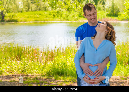 Pregnant woman and her husband in a park near the water hugging Stock Photo