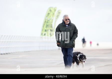 North Wales, UK Weather. The first named storm of season that brought 75mph gales to a central belt of the UK and North Wales now passing over North East of the UK. The Amber Warning for wind and rain is now reduce to yellow warning over North Wales as this dog walker discovered along the promenade with the blowing sands in Rhyl this morning, Denbighshire, Wales as this man discovered grimacing from the drifting sands Stock Photo