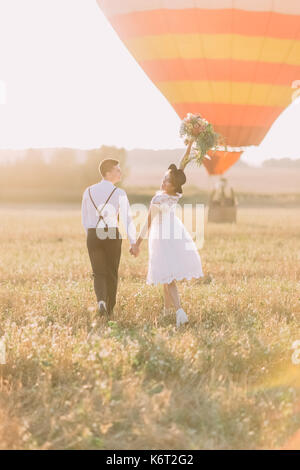 The happy newlyweds are walkong in the sunny field near the airballoon. The back view of the bride keeping up the bouquet of field flowers. Stock Photo