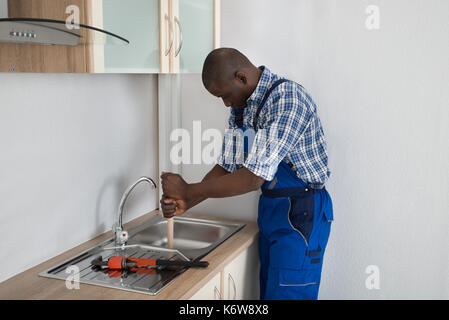 Young Male African Plumber Pressing Plunger In Kitchen Sink Stock Photo