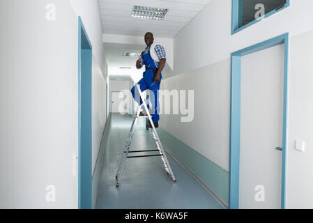Young African Male Electrician On Stepladder Gesturing Thumbs Up Sign Stock Photo