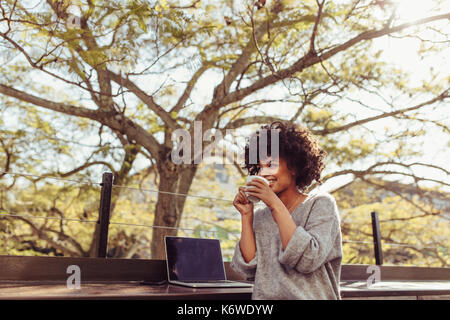 Curly haired woman sitting outdoors with coffee cup in her hands. Happy young woman sitting outdoors with a laptop and coffee. Stock Photo