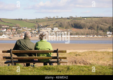 A couple sit on a bench facing the small village of Ferryside on the oposite shore in the distance and overlooking the river Towy, Carmarthenshire. Wa Stock Photo