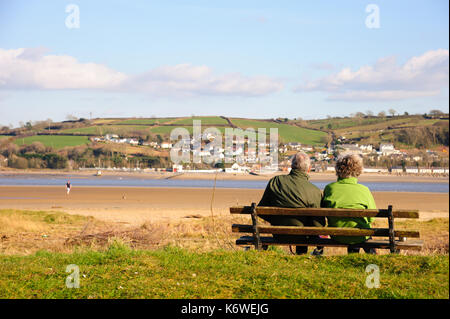 A couple sit on a bench facing the small village of Ferryside on the oposite shore in the distance and overlooking the river Towy, Carmarthenshire. Wa Stock Photo