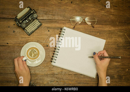 Woman hand writing in notebook, drinking coffee. Table with rustic wood texture. Stock Photo