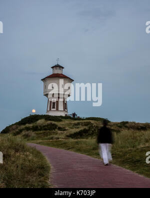 Wasserturm Langeoog. Germany Deutschland. Soon after twilight a moonrise occurs behind the water tower.  The water tower is a cultural landmark and major tourist attraction.  The clear sky allows the moon to be seen as it rises above the sand dune that the water tower was built upon.  A tourist sees the moonrise as she approaches the water tower on the footpath in the foreground.  This was photographed with a slow shutter speed to capture movement as the woman walks along the foot path. Stock Photo