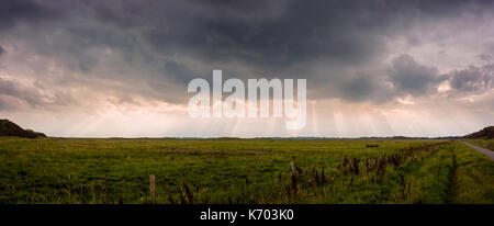 Melkhörndüne, Langeoog.  Deutschland.  Germany.  A panoramic skyscape with rays of sun shining though the threatening storm clouds.  Below lies the grassland of the national park between the sand dunes, Melkhörndüne.  The outline of the town is visible on the horizon. Stock Photo