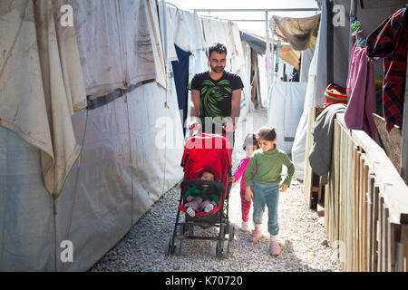 A father with two young girls at his side, pushes a stroller carrying the third child on the gravel path between refugee camp homes. Stock Photo